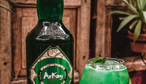 Green bottle of alcohol-free whisky next to glass with green drink garnished with cucumber and mint leaves.