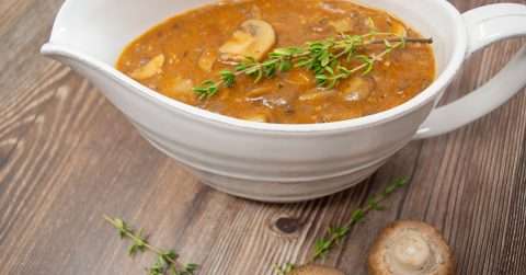 Gravy with mushrooms and thyme in white gravy bowl on a table with mushroom and thyme garnish.