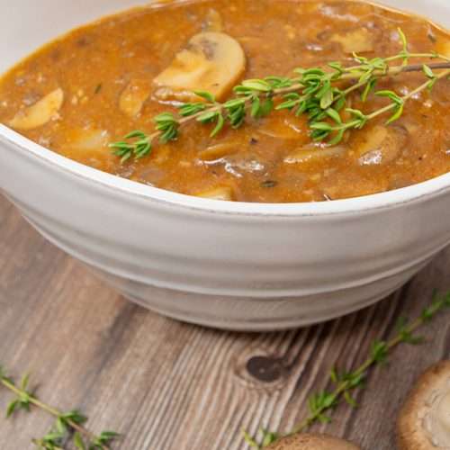 Gravy with mushrooms and thyme in white gravy bowl on a table with mushroom and thyme garnish.