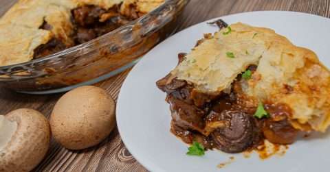 Pot pie with mushrooms on white plate surrounded by mushrooms and casserole dish of pie with slice missing.