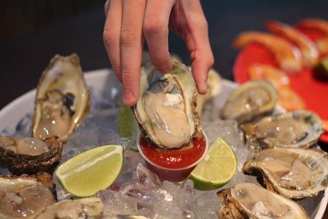 Hand dipping oyster into cocktail sauce on a plate with ice surrounded by other open oysters and lime wedges.