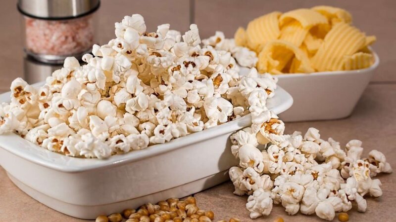 Bowl of popcorn and potato chips with popcorn spilling out onto a table.