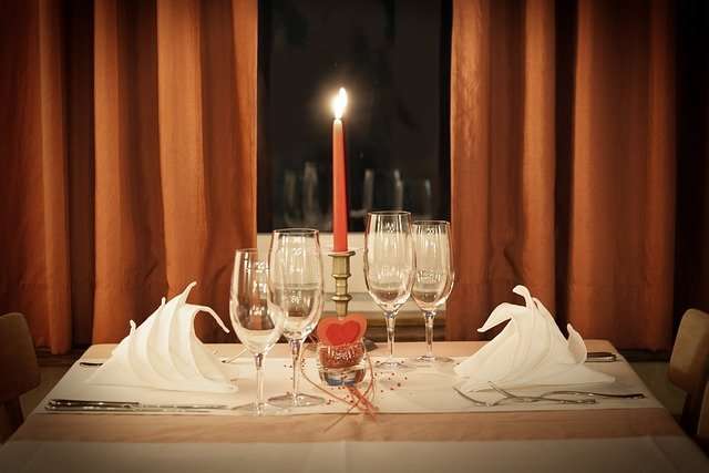 Red candle with heart at base surrounded with four whine glasses on a table with napkins and silverware.