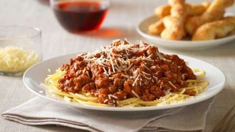 Bowl of pasta with meat sauce in a bowl with wine, cheese and bread on table.