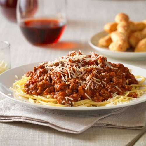 Bowl of pasta with meat sauce in a bowl with wine, cheese and bread on table.