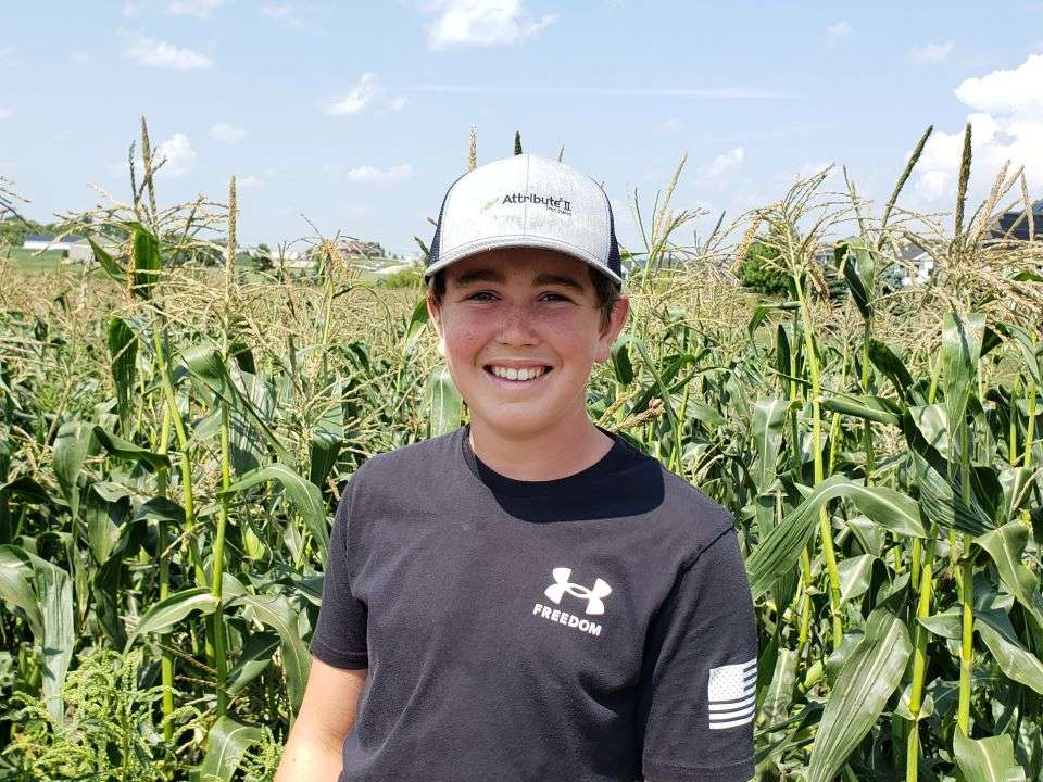 12-year-old Weston Hannan standing in front of his cornfield with blue shirt and baseball hat on.