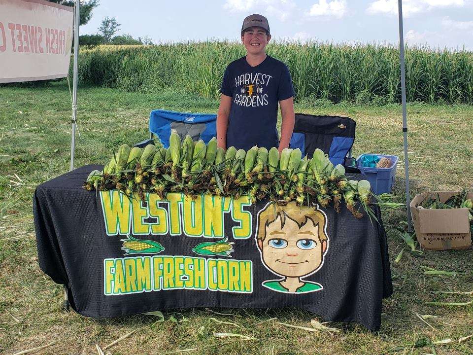 Weston Hannan in front of display of corn at the corn stand with cornfield in the background.