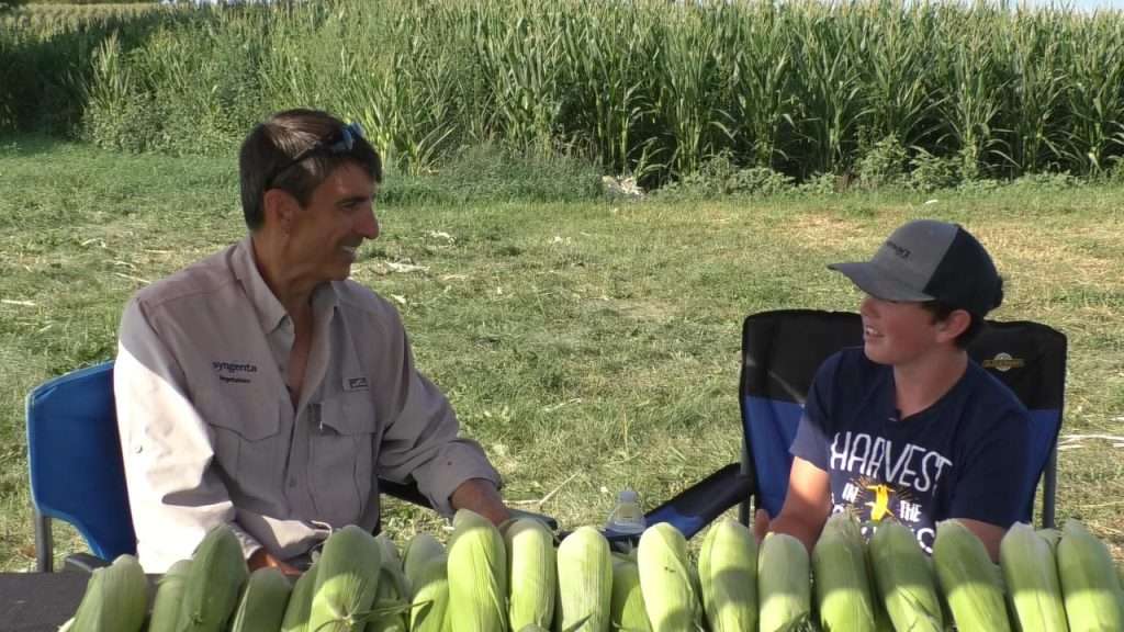 Glenn McKay and Weston Hannan talking at the corn stand in front of field of corn.