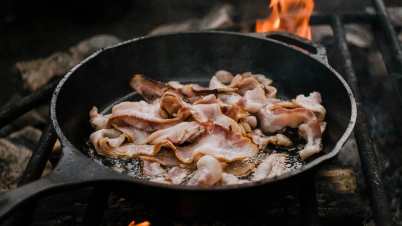 Cast iron pan with bacon cooking over a fire.