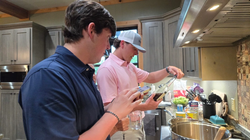 Luke holding cell phone while Jake measures wine in front of stove. 