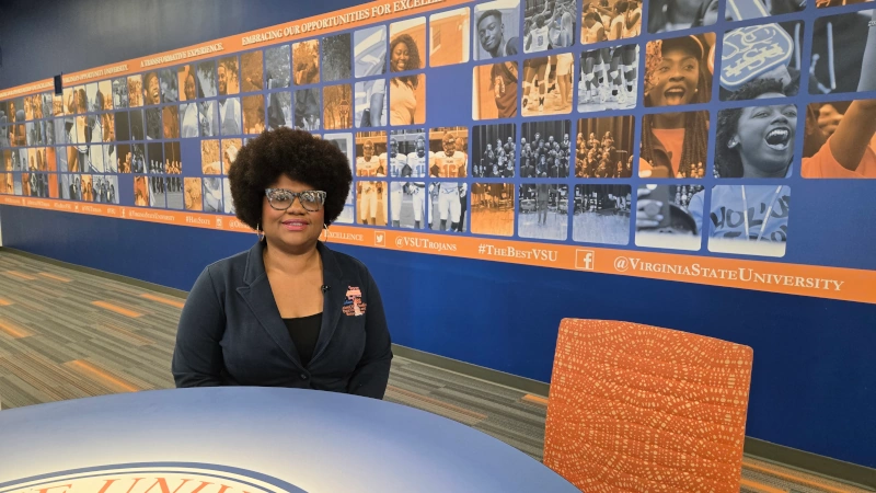 Dr. Cheryl Mango sitting at a table in front of Trojan mural in lobby of building.