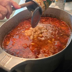 A step in the process of making pasta fazool, a woman adds a can of cannellini/navy beans to the pot.