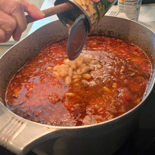 A step in the process of making pasta fazool, a woman adds a can of cannellini/navy beans to the pot.