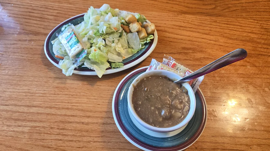A cup of soup with crackers and a side salad with croutons on a wooden table.